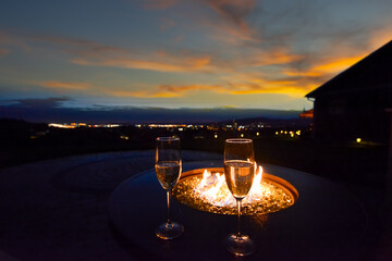 Two glasses of champagne placed on a flaming fire pit of a luxury hilltop home's back yard while overlooking a valley and city with lights illuminated at sunset in the evening.  - Powered by Adobe