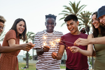 Happy young friends having fun holding sparkles at festival eve - Diverse group of multiracial people celebrating together on summer outdoors party