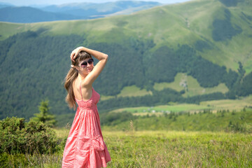 Young woman in red dress standing on grassy field on a windy day in summer mountains enjoying view of nature