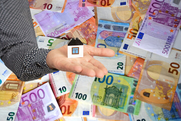 close-up of male hand holds model of house on lot of money background, paper euro banknotes, concept of cash, payments, savings, banking, save up to buy house, purchase real estate in mortgage, loan