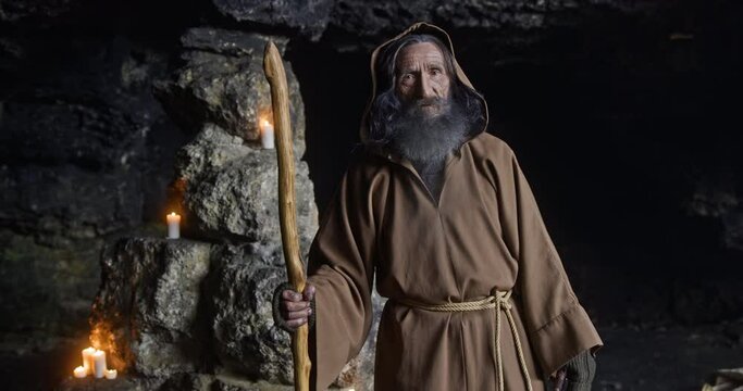 Medieval monk, with a staff in a hooded place, stands in a dark cave with glowing candles in the background.