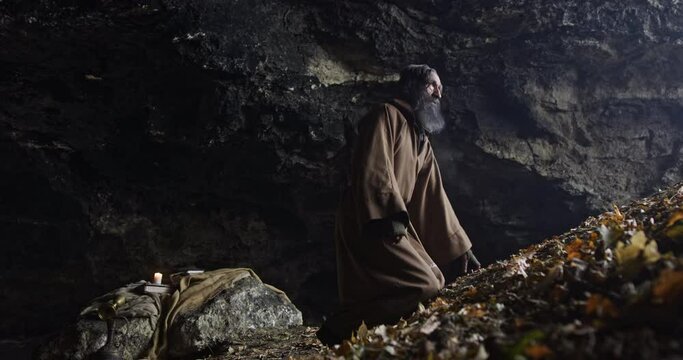 Hermit man with robe in the entrance of a old cave in front of a valley