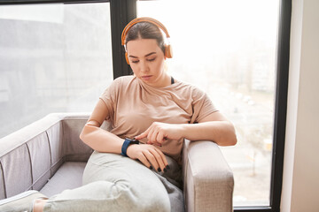 Female wearing headphones with arm implant sitting at the armchair and touching to the smartwatch