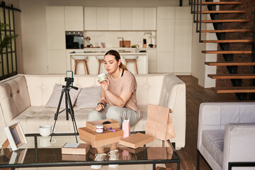 Female blogger influencer sitting at home and looking at cream while shooting herself on smartphone