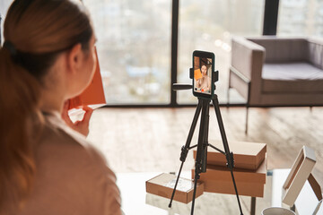 Woman blogger is sitting at the table and showing box with headphones to digital camera