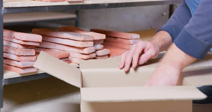Skilled worker takes handmade terracotta brick casts from wooden shelf putting into paper box. Decorative blocks for making tiles at workshop closeup