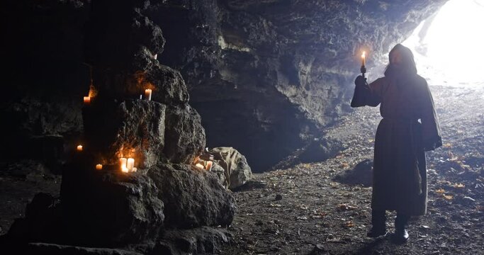 Friar with a candle in his hands wanders in a large dark cave on a background of lighted candles