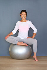 The body achieves what the mind believes. Full length shot of a sporty young woman sitting on a pilates ball against a grey background.