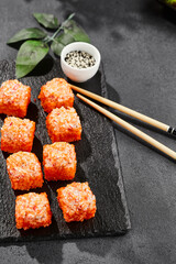Baked Maki sushi on dark slate. Hot california maki with crab. Sushi roll with masago outside, baked cheese and crab topped. Style concept japanese menu with black background, leaves and hard shadow.