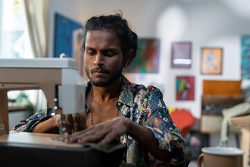 Serious stylish male tailor in non-gendered clothing creating new apparel while sitting in front of camera by electric sewing machine