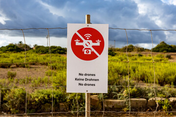 a white sign with a red prohibition sign saying "no drones". Sign on a fence indicating a no-fly zone. overcast gray sky. 