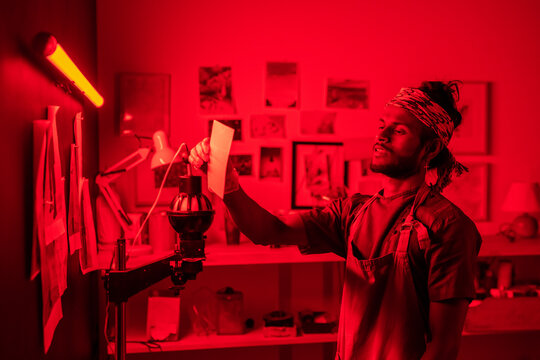 Happy young photographer looking at developed photo while holding it in front of himself in equipped red lit laboratory