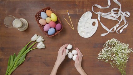 Top view of a table with items to create a composition for Easter. Women's hands put Easter eggs on...