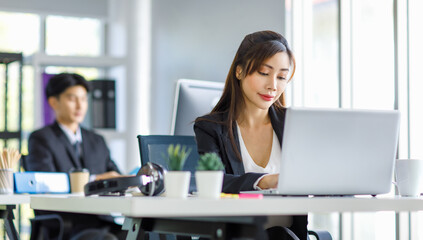 Obraz premium Asian young female professional businesswoman secretary employee in formal suit sitting at working desk typing report via laptop notebook computer while businessman manager work behind in office