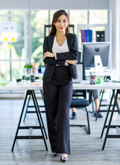 Asian young female professional businesswoman secretary employee staff in formal suit standing in front workstation desk crossed arms smiling in company office