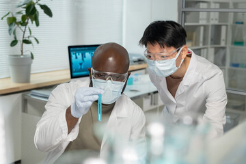 Two young interracial chemists in lab coats and protective masks looking at blue liquid in test...