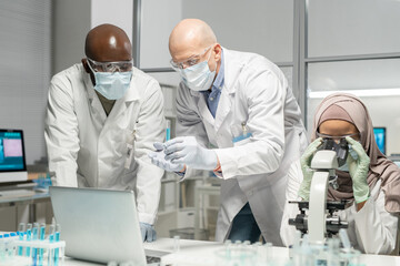 Mature clinician or scientist showing his African American colleague flask with blue liquid while Muslim female using microscope