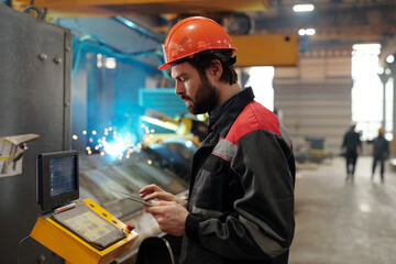 Side view of young engineer in workwear using digital tablet while standing in front of panel of huge industrial machine
