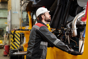 Young technician or repairman in hardhat and uniform standing by industrial machine while checking its engine system