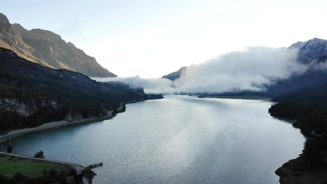 A sunrise aerial flyover up above Lake Sils in Maloja, Switzerland with a view of the peaks of Engadin and clouds above the water