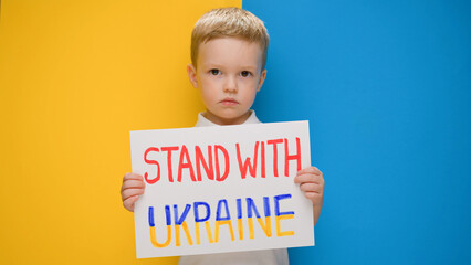 Portrait Blond little boy calls to Stand with Ukraine, raises banner with inscription stop the war in Ukraine standing on blue-yellow studio background. No war, stop war, russian aggression.