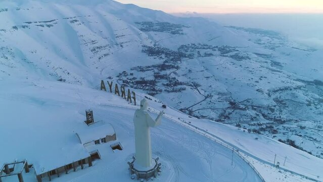 Drone rotation over Saint Charbel statue in snowy mountainous landscape at sunset. Faraya, Lebanon