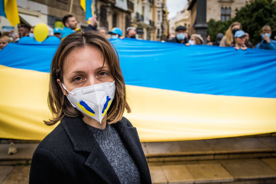 Sad Ukrainian Girl With Face Mask Painted in the Colors of Ukraine and a Large National Flag Behind Looking at the Camera