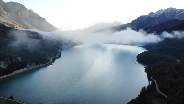 A sunrise aerial flyover through the clouds up above over Lake Sils in Maloja, Switzerland with a view of the peaks of Engadin and clouds moving over the water