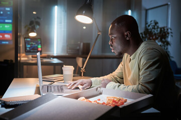 African businessman sitting at workplace and having a snack with pizza and coffee while working on laptop at office till late night - obrazy, fototapety, plakaty