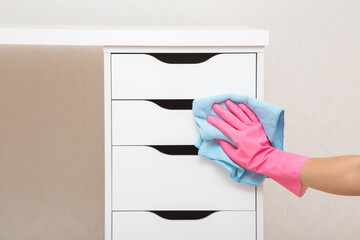 Young woman hand in pink rubber protective glove holding blue dry rag and wiping dust from white drawers of work table at home office. Closeup. General or regular cleanup in room. Cleaning service.