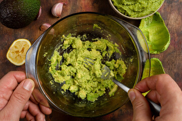 Man's hands mashing several avocados in a bowl with a fork to prepare a rich and fresh guacamole. Ingredients in composition on a wooden background in brown tones. Healthy food concept