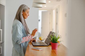 Female person standing by wooden table and holding cup of coffee while using laptop in kitchen