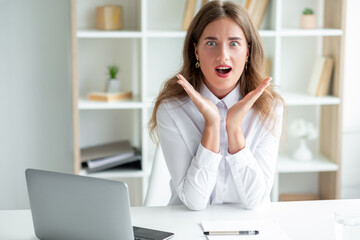 Pleasant news. Excited woman. Office work. Surprised pretty lady sitting desk with opened laptop holding hands up looking impressed in light room interior. - obrazy, fototapety, plakaty