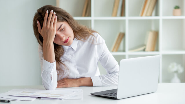 Work trouble. Tired woman. Online conference. Displeased clueless lady holding hand on forehead sitting desk with documents and opened laptop in light room interior.
