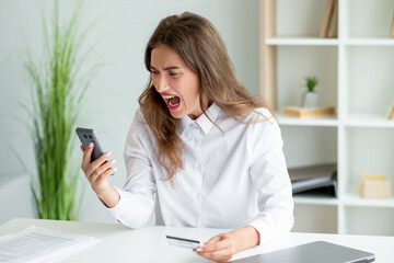 Payment problem. Furious woman. Mobile application. Shouting mad office lady holding credit card looking smartphone sitting work desk in light room interior.