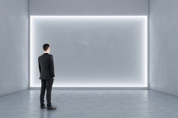Fototapeta premium Businessman standing in modern exhibition hall interior with illuminated white mock up wall and concrete floor. Gallery concept.