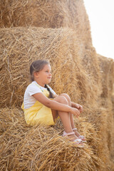 Naklejka na ściany i meble Pretty thinking female kid resting on hayrick with slight smile on face looking away and holding hay in hands wearing sundress. Having fun away from city on field full of golden hay.