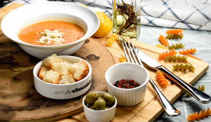 Tomato soup garnished with pepper, parsley and  parmesan cheese in white bowl on wooden board with bread cube and red black pepper and tricolor fusilli on side