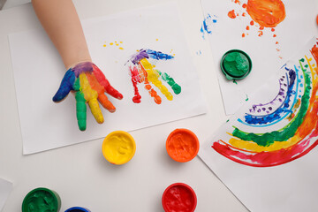 Little child making hand print on paper with painted palm at white table indoors, top view