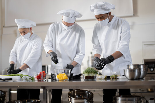 Three well dressed chefs in face masks prepare takeaway food in professional kitchen. Concept of a dark kitchen for cooking for delivery during pandemic