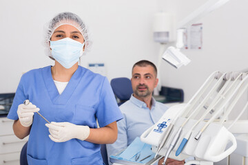 A portrait of a handsome female dentist in face mask with patient in the background at dental clinic