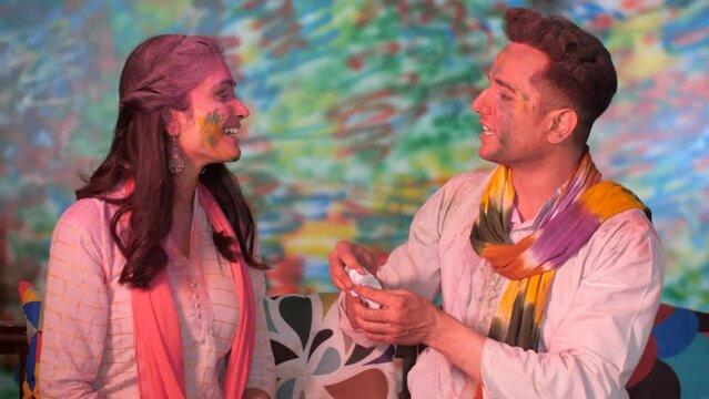 A man wiping off the Gulal on his friend's pretty face using a tissue paper - Holi festival celebration  organic colors. A young couple spend time together at home during the Indian festival of col...