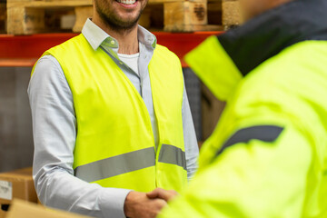 wholesale, logistic, people, agreement and export concept - close up of manual worker and businessmen in reflective safety vests shaking hands and making deal at warehouse