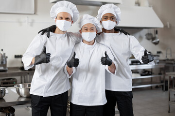Portrait of multiracial team of three chefs standing together and showing OK sign. Well-dressed chefs wearing face masks and protective gloves to prevent infection spread during pandemic