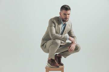 sexy man in suit looking away while crouching on wooden chair