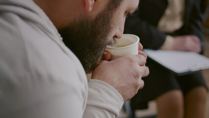 Close up of man drinking cup of coffee and shaking at aa group therapy session. Adult holding hot beverage while trembling because of alcohol addiction, attending rehab program for recovery.
