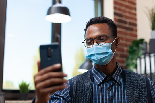 Portrait of startup office worker with glasses wearing face mask holding smartphone in online interview. Focus on entrepreneur in isolation in video call looking at mobile phone screen. - Powered by Adobe