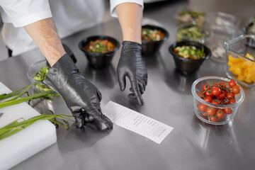 Chef pointing on printed check with order, cooking food for delivery in professional kitchen. Close-up on table with food ingredients. Dark kitchen and take away food concept