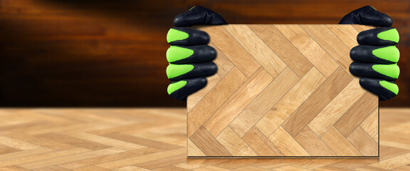 Two hands with protective work gloves holding a herringbone wood parquet sample, on top of a similar floor, with copy space.
