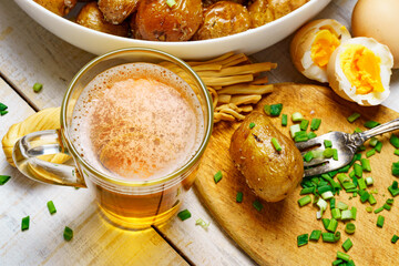 tasty food in rural style, - glass of beer, fried chicken meat and potatoes, garlic and green onions, cheese, boiled eggs and salt, cooked food on a white wooden boards